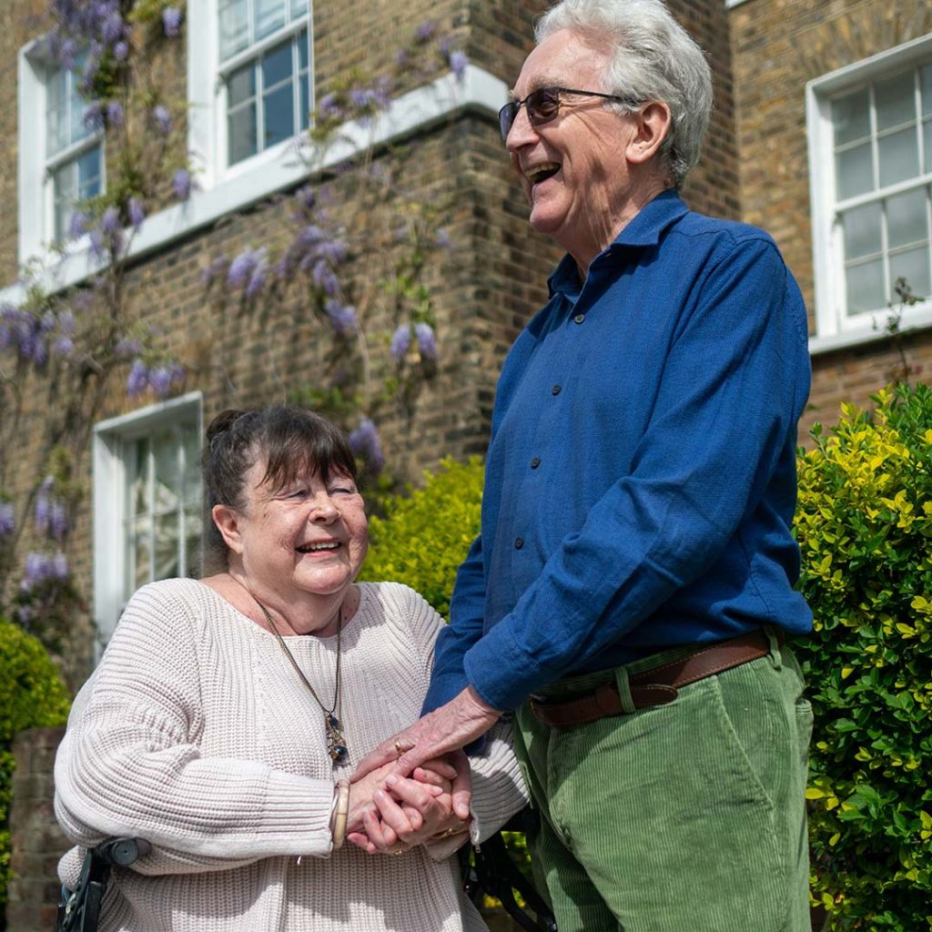 A man with gray hair holds the hand of a smiling woman in a wheelchair